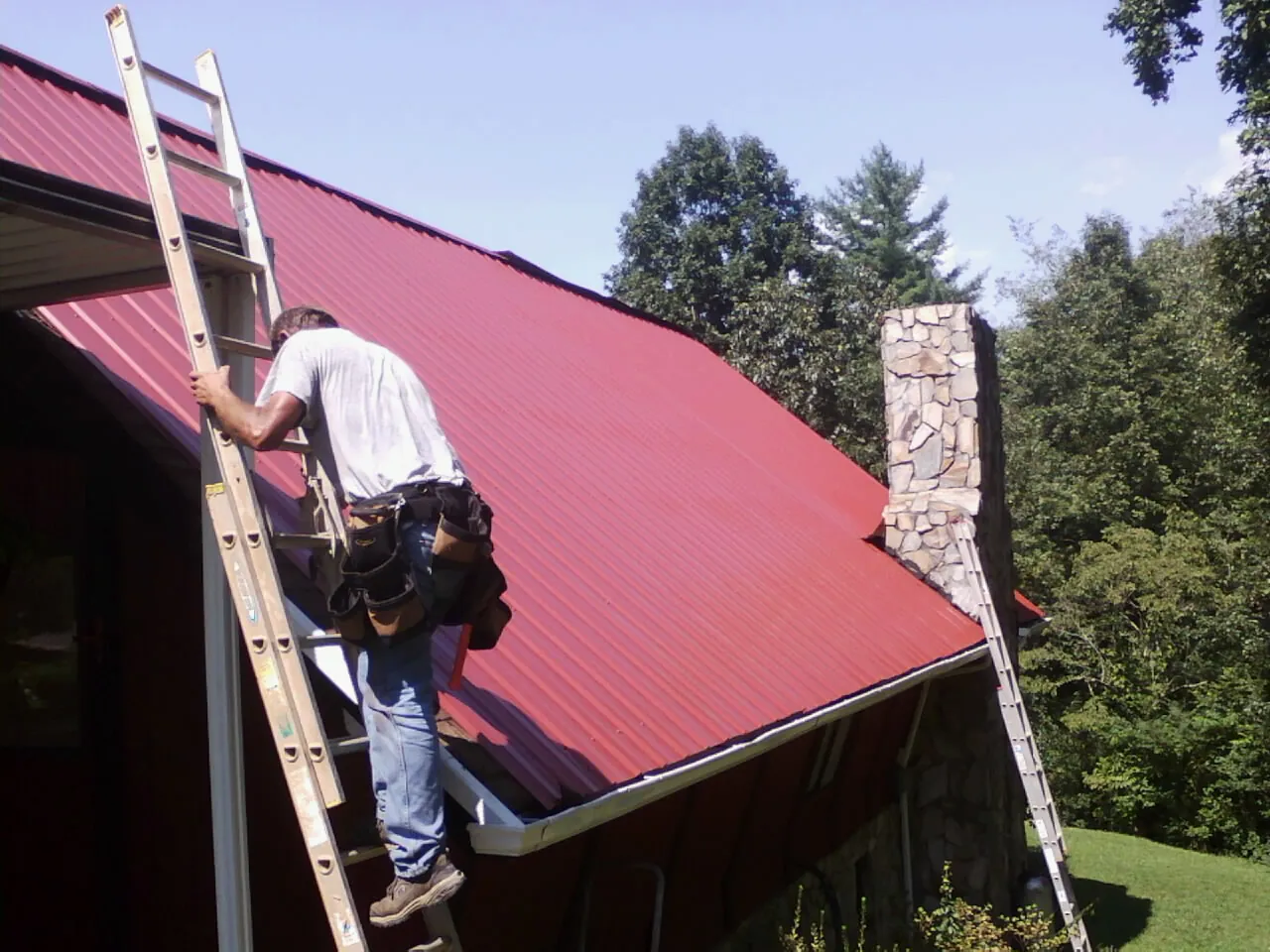 Roofer on a ladder beside a red metal roof and stone chimney — Soffit & Fascia Repair by Roofing Brothers in Montrose